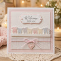 Pink baby card with pastel clothing border, twine bow, and Welcome Little One greeting