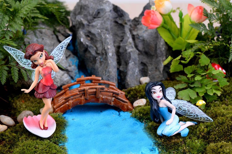 Two fairy figurines with wings, one in red and one in blue, are placed in a miniature garden setting with a small wooden bridge, a stream, and green moss.
