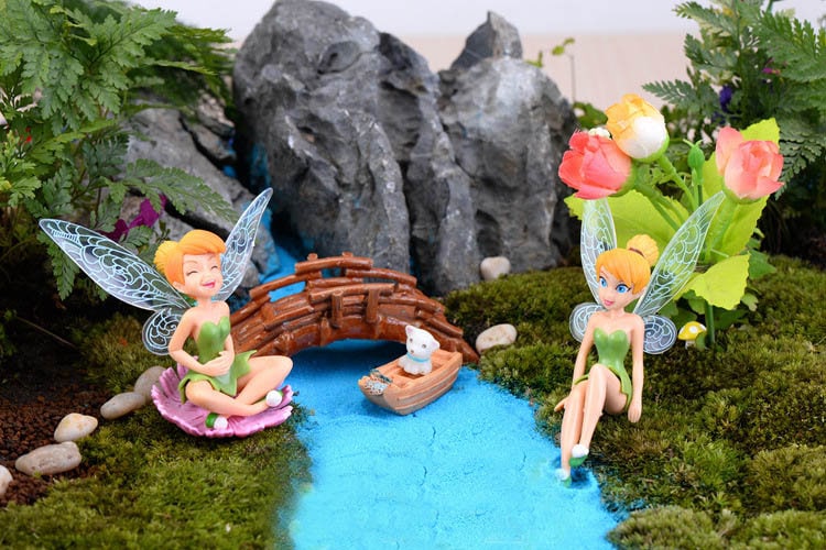 Two fairy figurines with translucent wings, one sitting on a pink flower and the other sitting on a rock by a blue stream. A small wooden bridge and a white boat with a cat figurine are in the background.