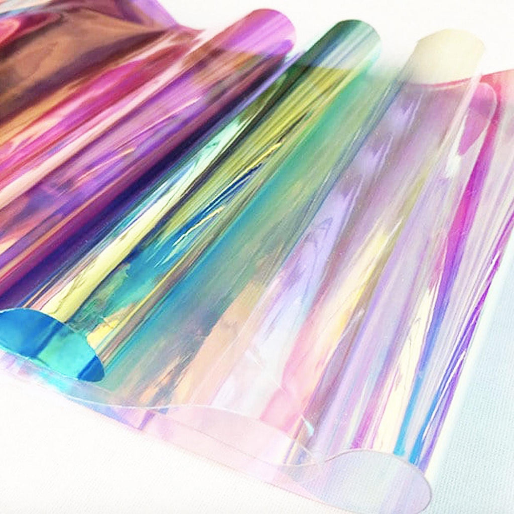 Several rolls of iridescent cellophane paper. The paper reflects a spectrum of colors, including pink, purple, green, and blue. The rolls are tightly wound, and the edges are visible, creating a layered effect. The overall aesthetic is shiny and ethereal.