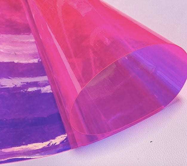 A sheet of iridescent, translucent material in shades of pink and purple. The material is rolled up, revealing a layered effect of colors. The surface reflects light, creating a shimmering appearance.